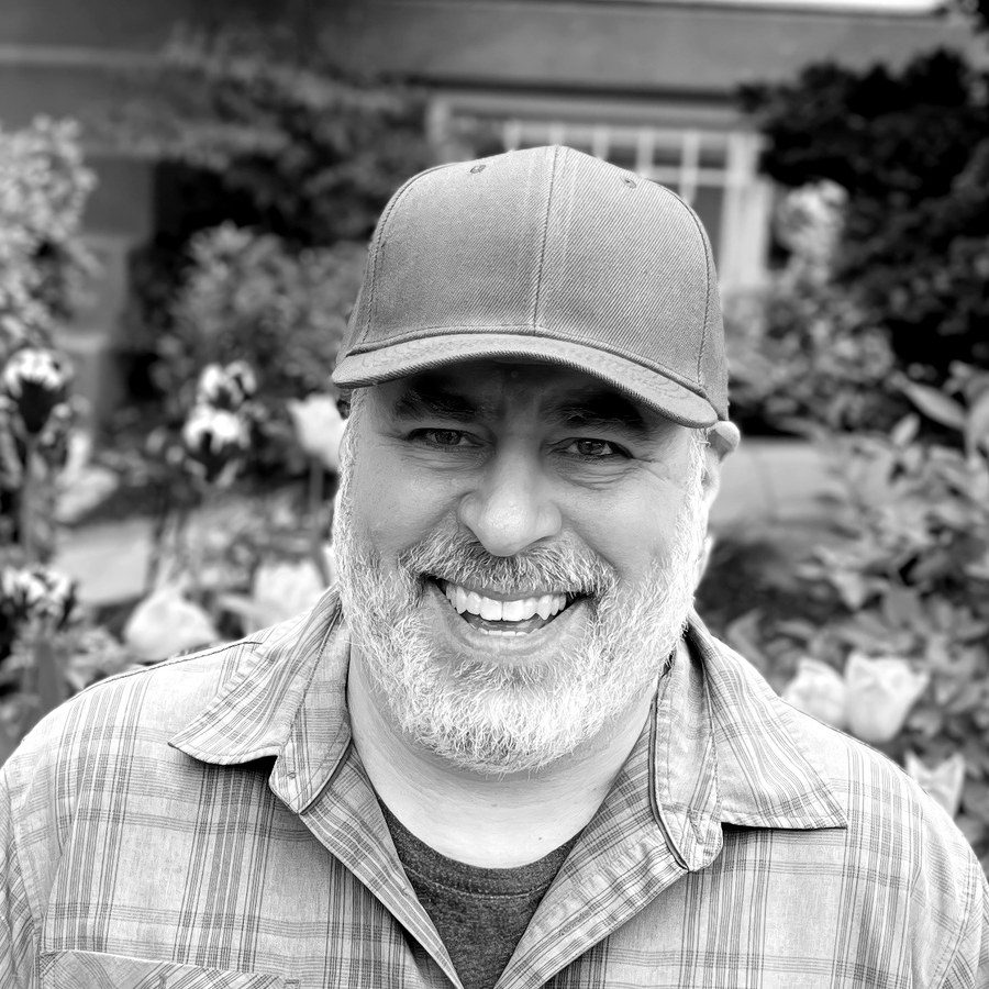 Black-and-white portrait of Doug Dworkin smiling in a garden, wearing a ball cap and plaid shirt.