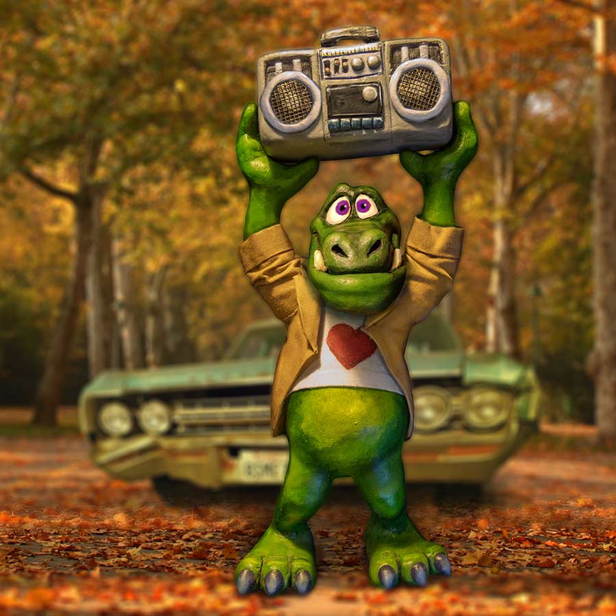 Photograph of a hand-sculpted green monster in a tan jacket and heart t-shirt holding a boombox overhead, composited onto an autumn street with a vintage car behind him.